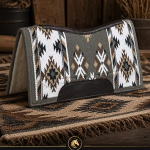 Op de afbeelding: Een zadeldek met een geometrisch patroon in grijs, wit, zwart en bruin, voorzien van donkerbruine lederen accenten langs de rug en zijkanten. De onderzijde is gevoerd met een dikke, crèmekleurige fleece en de randen zijn afgewerkt met grijze bies.