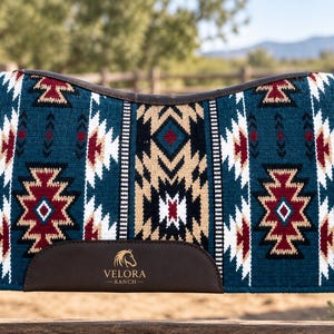 May include: A rectangular horse saddle pad featuring a teal, tan, white, and maroon geometric pattern. The pad has dark brown leather wear guards at the bottom, with the gold text VELORA RANCH and a horse head logo printed on the centre guard.