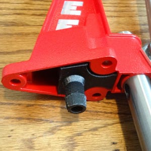 May include: A bright red metal tool component featuring a textured finish and a black cylindrical nozzle attachment. The structure includes a silver metallic arm extending to the right, secured by a visible bolt, and white lettering partially visible.