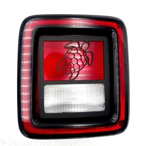 May include: A square, black and red vehicle tail light with a turtle design. The tail light has a red border and a red center with a black turtle silhouette. The bottom section is white with a grid pattern.