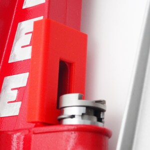 May include: A red metal reloading press component featuring a bright red vertical frame with white lettering that reads LEE. A small, rectangular red plastic attachment is mounted to the frame, positioned above a circular silver metal shell holder.