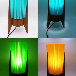 May include: A table lamp featuring a cylindrical, vertically ribbed shade in turquoise, green, or yellow, supported by a brown, three-legged stand. The lamp includes a black power cord and emits a bright, colored glow when illuminated.