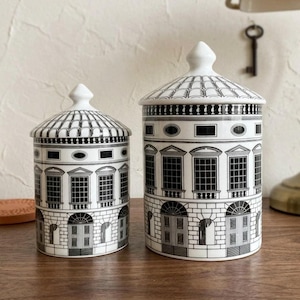 May include: Two cylindrical ceramic canisters with lids, featuring a black and white architectural illustration of a building facade with arched doorways, grid-patterned windows, and a tiled roof design. Both containers have a matching finial.