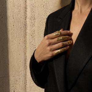 May include: A set of four gold rings with organic, textured surfaces worn on the fingers. The rings feature irregular, shell-like shapes that cover a portion of each finger. The jewellery is displayed against a black blazer with a lapel.