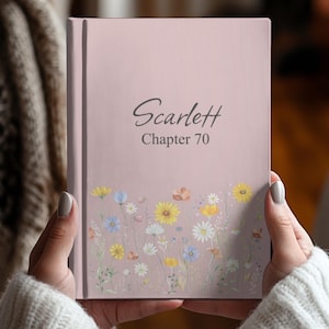 May include: A light pink hardback book featuring the text Scarlett Chapter 70 in a dark grey script and print font. The lower portion of the cover is decorated with a colourful illustration of various wildflowers in shades of yellow, white, and blue.