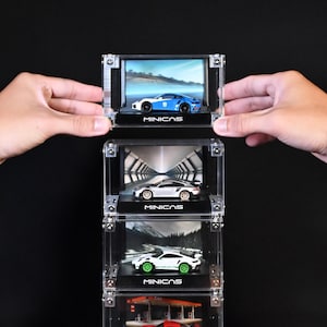 Może przedstawiać: Stos czterech przezroczystych akrylowych gablot, z których każda zawiera miniaturowy model samochodu sportowego i unikalne tło. Podstawa każdej gabloty zawiera napis MINICAS białymi literami. Górna gablota zawiera niebiesko-biały samochód.