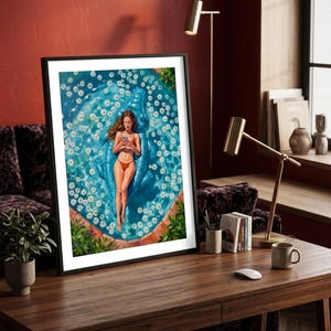 May include: A framed wall art print depicting a person in a red bikini lying on a blue crocodile-shaped pool float. The float is surrounded by white daisy flowers in a turquoise swimming pool with terracotta-colored edges and green foliage.