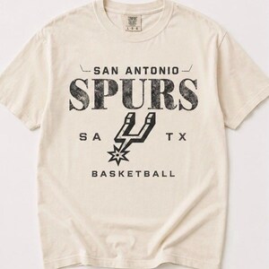 May include: A cream-colored short-sleeve t-shirt featuring black distressed text that reads SAN ANTONIO SPURS, SA TX, and BASKETBALL. The center graphic displays the San Antonio Spurs basketball team logo in black. The shirt has a crew neckline.