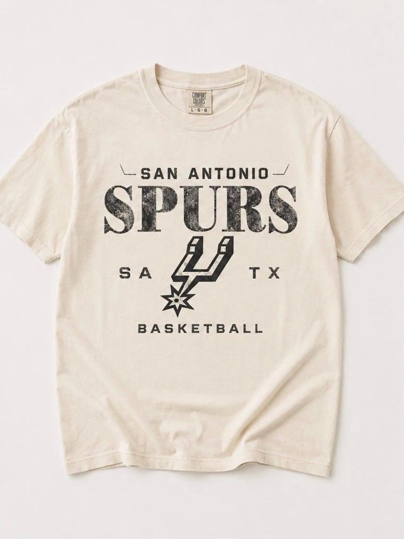 May include: A cream-colored short-sleeve t-shirt featuring black distressed text that reads SAN ANTONIO SPURS, SA TX, and BASKETBALL. The center graphic displays the San Antonio Spurs basketball team logo in black. The shirt has a crew neckline.