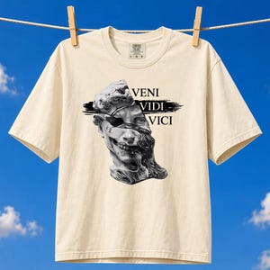 Könnte beinhalten: Ein cremefarbenes Kurzarm-T-Shirt mit einer schwarz-weißen Grafik eines klassischen Statuenkopfes mit Augenklappe und dem in Schwarz gedruckten Text VENI VIDI VICI. Das T-Shirt hat einen Rundhalsausschnitt und kurze Ärmel.