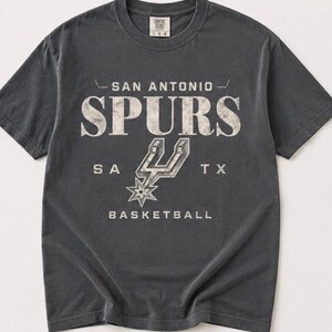 May include: A charcoal gray short-sleeve t-shirt featuring the text SAN ANTONIO SPURS, SA TX, and BASKETBALL in a distressed white font. The graphic includes the San Antonio Spurs logo in the center. The shirt has a crew neckline and a tag.