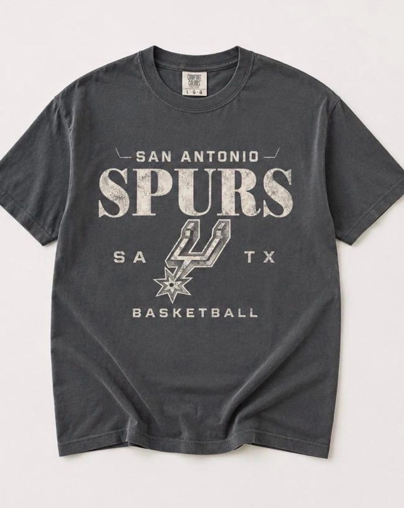 May include: A charcoal gray short-sleeve t-shirt featuring the text SAN ANTONIO SPURS, SA TX, and BASKETBALL in a distressed white font. The graphic includes the San Antonio Spurs logo in the center. The shirt has a crew neckline and a tag.