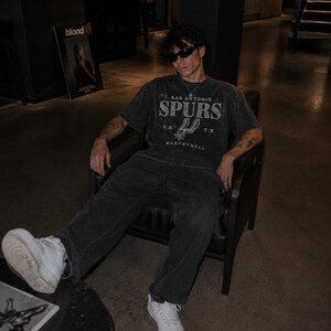 May include: A charcoal gray short-sleeve t-shirt featuring the text San Antonio Spurs and the team logo in a distressed white print. The shirt is paired with dark gray denim jeans and white sneakers, creating a casual outfit with a vintage aesthetic.