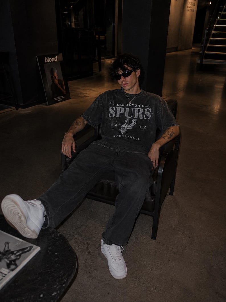 May include: A charcoal gray short-sleeve t-shirt featuring the text San Antonio Spurs and the team logo in a distressed white print. The shirt is paired with dark gray denim jeans and white sneakers, creating a casual outfit with a vintage aesthetic.