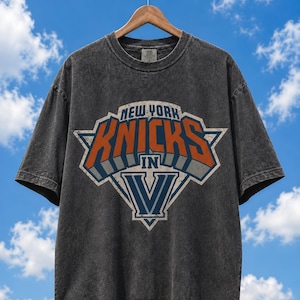 Op de afbeelding: Een antracietgrijs t-shirt met korte mouwen en een grafische print met de tekst NEW YORK KNICKS IN V. Het ontwerp bevat oranje, blauwe en witte letters in een stervormig embleem. De stof heeft een gewassen, vintage uitstraling.