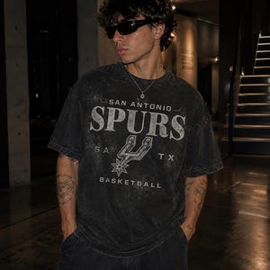 May include: A charcoal grey, acid-washed short-sleeve t-shirt featuring the text SAN ANTONIO SPURS BASKETBALL and the team logo printed in a distressed white font. The shirt has a crew neckline and a relaxed fit, showcasing a vintage-inspired aesthetic.