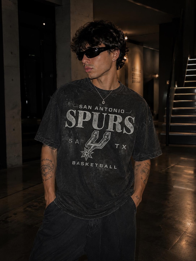 May include: A charcoal grey, acid-washed short-sleeve t-shirt featuring the text SAN ANTONIO SPURS BASKETBALL and the team logo printed in a distressed white font. The shirt has a crew neckline and a relaxed fit, showcasing a vintage-inspired aesthetic.