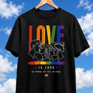 Könnte beinhalten: Ein schwarzes T-Shirt mit einer Grafik, die das Wort LOVE in Regenbogenfarben über einer weißen Strichzeichnung von vier Gesichtern zeigt. Darunter befindet sich ein Regenbogen-Pinselstrich, der Text LOVE IS LOVE und BE PROUD. BE YOU. BE FREE.