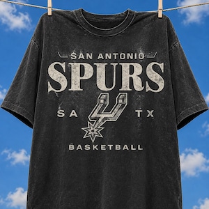 May include: A black, short-sleeved t-shirt with a distressed, vintage-style graphic. The print features the text "SAN ANTONIO SPURS" in large, weathered white letters, the team's spur logo, the letters "SA TX", and the word "BASKETBALL" below.