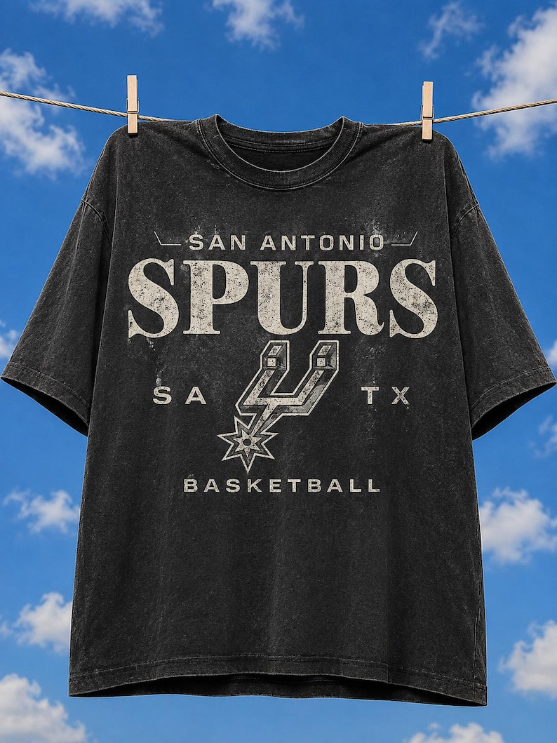 May include: A black, short-sleeved t-shirt with a distressed, vintage-style graphic. The print features the text "SAN ANTONIO SPURS" in large, weathered white letters, the team's spur logo, the letters "SA TX", and the word "BASKETBALL" below.