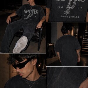 May include: A charcoal gray short-sleeve t-shirt featuring a distressed graphic print that reads SAN ANTONIO SPURS with the team logo, SA, and TX. The shirt has a crew neckline and a washed-out, vintage-inspired texture throughout the fabric.
