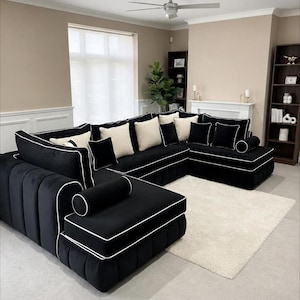 May include: A black velvet U-shaped sofa with white trim and cushions. The sofa is in a living room with a cream rug, neutral walls, and a ceiling fan. The sofa has a modern design and is a statement piece.