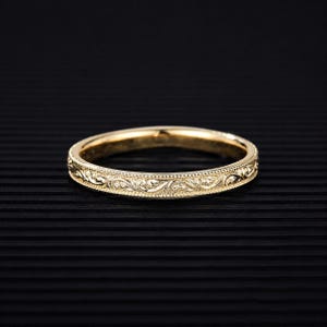 May include: A gold band ring featuring an intricate engraved scrollwork pattern along the outer surface. The edges of the ring are finished with a fine beaded milgrain texture, creating a detailed and polished appearance on the metallic surface.