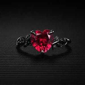 May include: A black ring featuring a large, heart-shaped, faceted red gemstone. The ring has a dark, ornate band with leaf and small black stone accents. The ring is set against a black background.