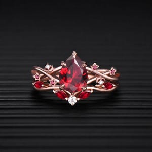 May include: A rose gold ring featuring a large pear-shaped red centre stone, accented with smaller marquise-cut red stones, tiny round white stones, and star and crescent moon motifs integrated into the band, creating a celestial-inspired design.