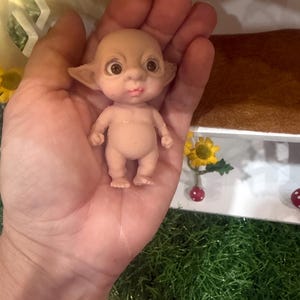 May include: A small, pale-skinned figurine with a bald head, large dark eyes, and pointed ears. The creature has a rounded torso, short limbs, and a small mouth with pinkish lips. It is depicted in a standing position with a neutral facial expression.