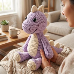 Può includere: Un giocattolo di dinosauro all'uncinetto fatto a mano nei toni del lavanda e crema. Il peluche ha grandi occhi neri, una bocca sorridente e una pancia e spine color crema. Il dinosauro è seduto su una coperta di maglia.