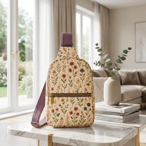 May include: A cream-colored sling backpack featuring a repeating pattern of small wildflowers in shades of red, yellow, and blue. The bag includes a solid purple adjustable strap, a top handle, and a horizontal front zipper pocket with gold hardware.