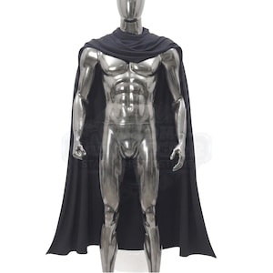 May include: A solid black cape featuring a draped cowl neckline that rests around the shoulders and extends down the back. The garment is displayed on a metallic silver mannequin, showcasing the smooth fabric texture and the full-length design.