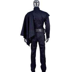 May include: A dark navy blue long-sleeved jumpsuit worn by a mannequin, featuring a high collar, a structured shoulder panel, a matching dark cape draped over the right shoulder, and a wide black utility belt secured around the waist area.