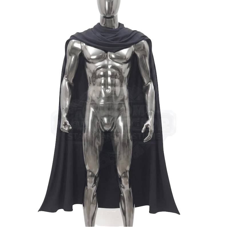 May include: A solid black cape featuring a draped cowl neckline that rests around the shoulders and extends down the back. The garment is displayed on a metallic silver mannequin, showcasing the smooth fabric texture and the full-length design.