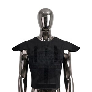 Peut inclure: Un gilet tactique noir doté de protections d'épaules et de bandes auto-agrippantes verticales sur la poitrine. Le vêtement est présenté sur un mannequin métallique, arborant une coupe structurée avec une encolure ronde et des manches courtes.