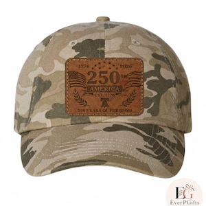Può includere: Cappellino da baseball mimetico con toppa in pelle marrone. La toppa presenta la scritta "250th America Est. 1776" con bandiere americane e gli anni 1776 e 2026. Il cappellino è di colore neutro.