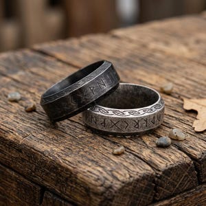 Op de afbeelding: Twee metalen ringen met een donkere, verweerde afwerking. Elke band is voorzien van gegraveerde runentekens in het midden en een decoratief vlechtpatroon langs de boven- en onderrand. De ringen zijn cirkelvormig met een plat profiel.