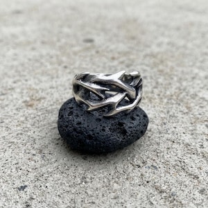 May include: A silver-toned metal ring featuring an intricate, interconnected design of sharp, thorn-like branches. The band has a weathered, oxidised finish with deep crevices between the pointed elements, creating a textured and dimensional appearance.