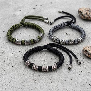 Könnte beinhalten: Drei geflochtene Kordelarmbänder in Olivgrün, Anthrazit und Schwarz. Jedes Armband ist mit vier zylindrischen, silberfarbenen Metallperlen versehen, die mit Runensymbolen graviert sind. Die Armbänder verfügen über verstellbare Kordelverschlüsse.