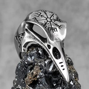 May include: A silver-toned metal ring shaped like a bird skull, featuring an engraved Vegvisir compass symbol on the cranium and a triangular Valknut emblem on the side. The surface has a polished finish with darkened recessed details.