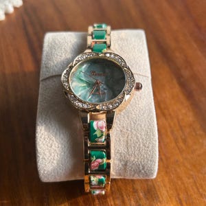 Può includere: Un orologio da polso color oro con un cinturino verde e a fantasia floreale. Il quadrante dell'orologio è circondato da un anello di cristalli trasparenti. Il quadrante ha un design marmorizzato verde e bianco.