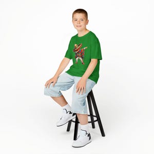 Pode incluir: Uma camiseta verde com um gráfico de uma pessoa com um chapéu e poncho fazendo o dab. A pessoa está usando um poncho colorido, um chapéu marrom e calças brancas. A pessoa está sentada em um banquinho preto, usando shorts azul claro e tênis brancos.