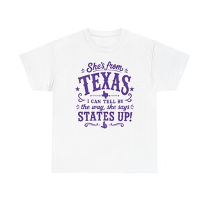 Könnte beinhalten: Ein weißes kurzärmeliges T-Shirt mit violettem Text, der lautet: She's from TEXAS I CAN TELL BY the way, she says STATES UP! Das Design enthält eine kleine Silhouette des Bundesstaates Texas sowie dekorative Schnörkel und Sterne.