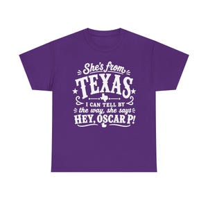 May include: A purple short-sleeved t-shirt featuring white text that reads: She's from TEXAS I CAN TELL BY the way, she says HEY, OSCAR P! The design includes a small graphic of the state of Texas and a small duck icon below the text.
