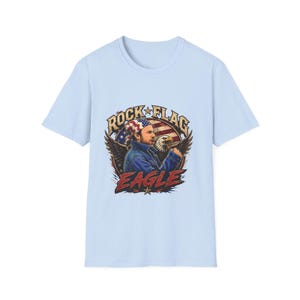 Puede incluir: Camiseta azul claro con un diseño gráfico de un hombre con un águila, la bandera estadounidense y las palabras "ROCK FLAG EAGLE". El diseño es de estilo vintage y patriótico.