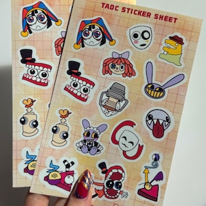 May include: Two rectangular sticker sheets labeled TADC STICKER SHEET at the top, featuring various cartoon character illustrations including a jester, a mask, a tooth character with a top hat, and other colorful figures on a grid background.