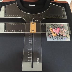 Può includere: Uno strumento di allineamento per magliette in acrilico trasparente con segni in centimetri è posto su una maglietta nera. Lo strumento ha un righello e un design a forma di T. È visibile un disegno a trasferimento termico con la parola "LOVE".