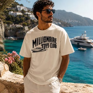 May include: An off-white short-sleeve t-shirt featuring a black graphic print on the chest. The design includes the text "MILLIONAIRE TOYS CLUB" in a bold, arched font above a stylized illustration of a sleek motorboat moving across water.