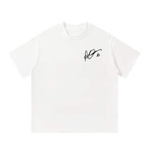 Pode incluir: Uma t-shirt branca de manga curta com gola redonda e um gráfico de assinatura manuscrita em preto com um pequeno símbolo de coração impresso na parte superior direita do peito. A peça é de cor branca lisa com um design simples.